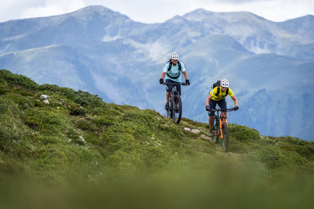 Zwei Mountainbiker fahren auf einem Bergpfad vor Bergkulisse