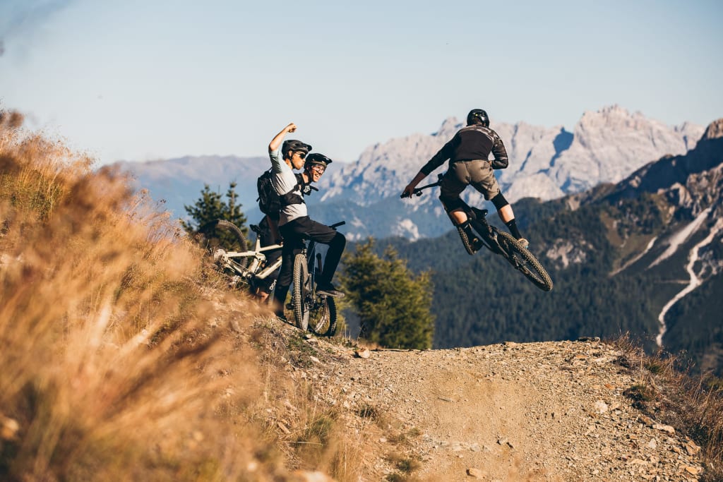 Mountain biker jumping on trail while friends cheer nearby in mountains